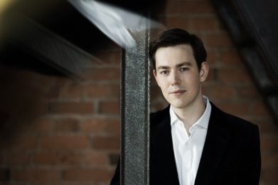 Man wearing a white shirt and black jacket stands against a metal beam. He is musician Edward Picton-Tuberbvill.