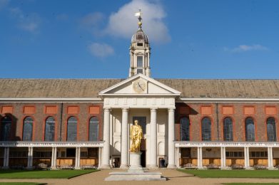 Galleried building with portico and tower. In front of the building is a gold statue.