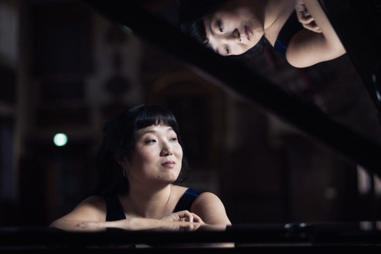 Woman seated at and leaning on a piano with her reflection on the piano lid.