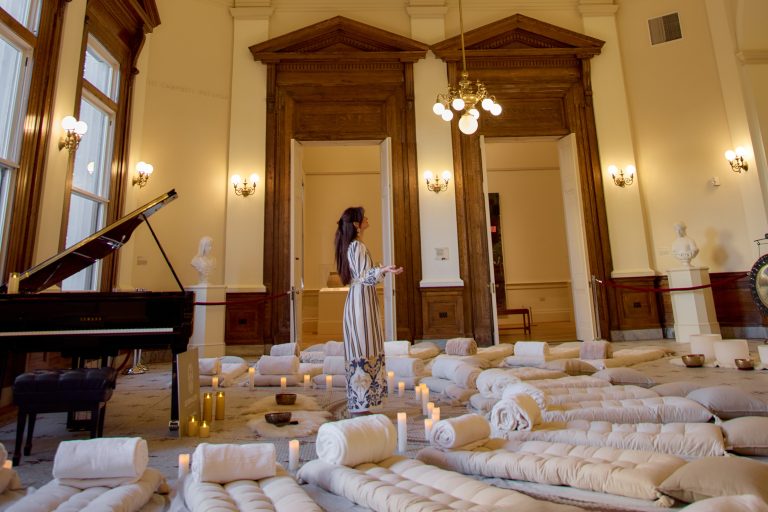 Woman stands in a room surrounded by yoga blankets and pillows. A piano is behind her.