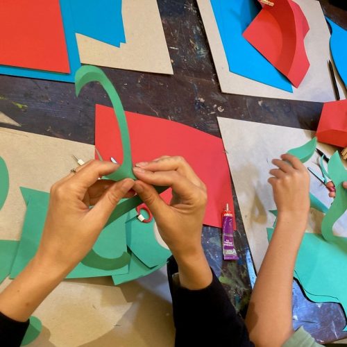 Image shows hand bending and folding coloured pieces of card. The table has card on it.
