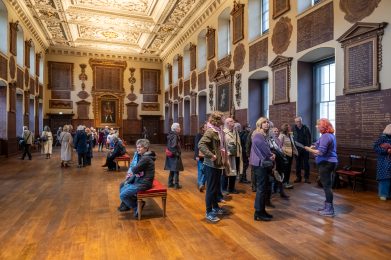 Group of people stood in a large room listening to a woman giving a tour.