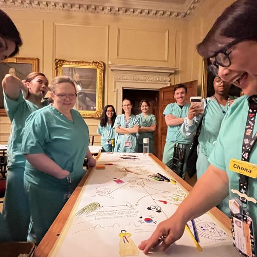 Group of people wearing healthcare scrubs stood pointing at a large scroll of paper with illustrations on it.