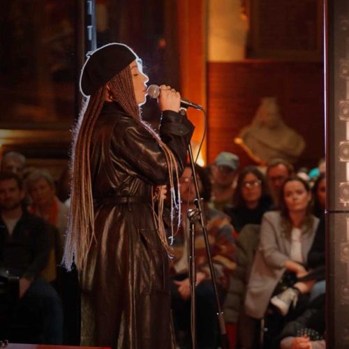 A singer wearing a leather coat and a beret performs in Barts Great Hall